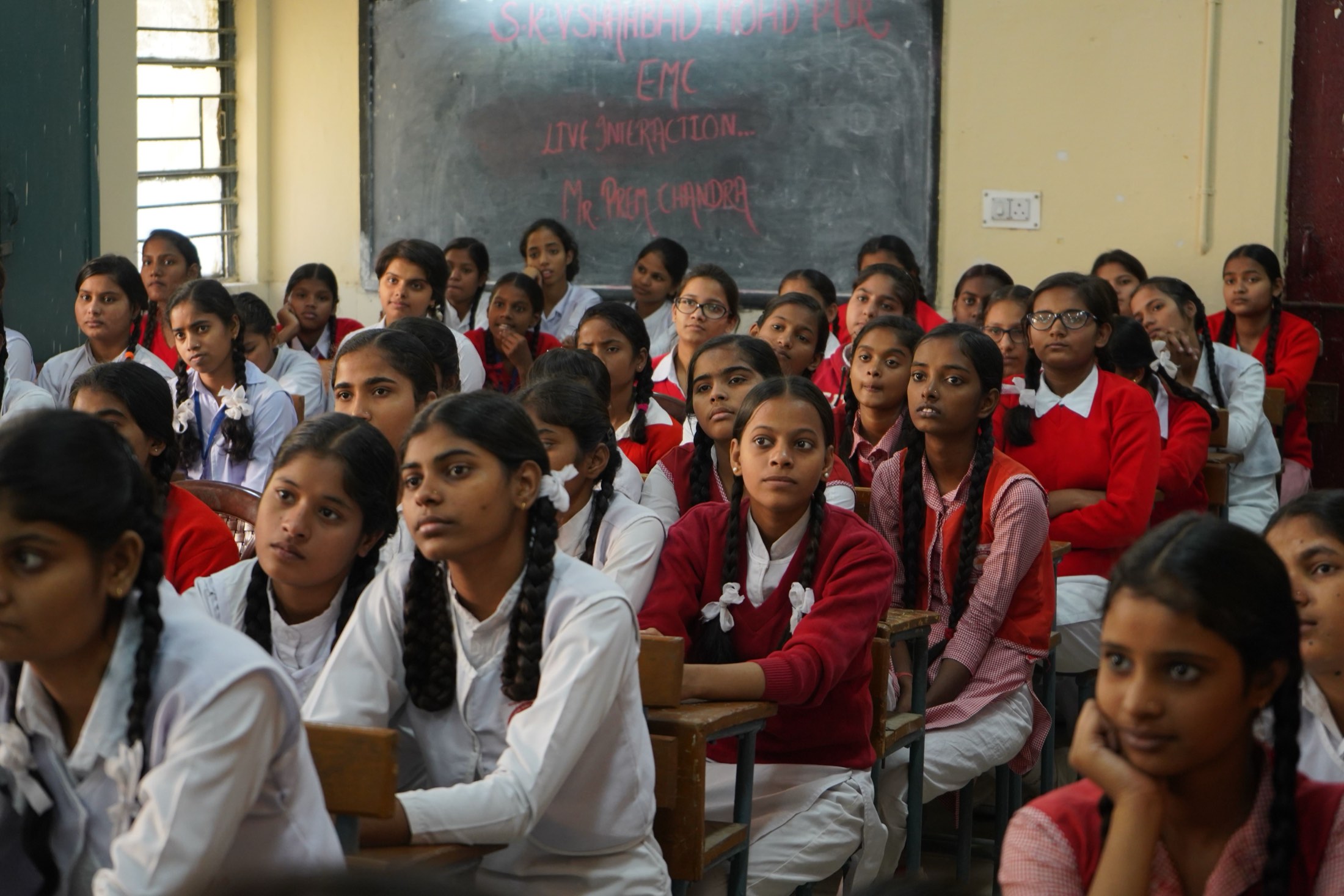 A classroom of schoolgirls in white and red uniforms seated at wooden desks, listening attentively.