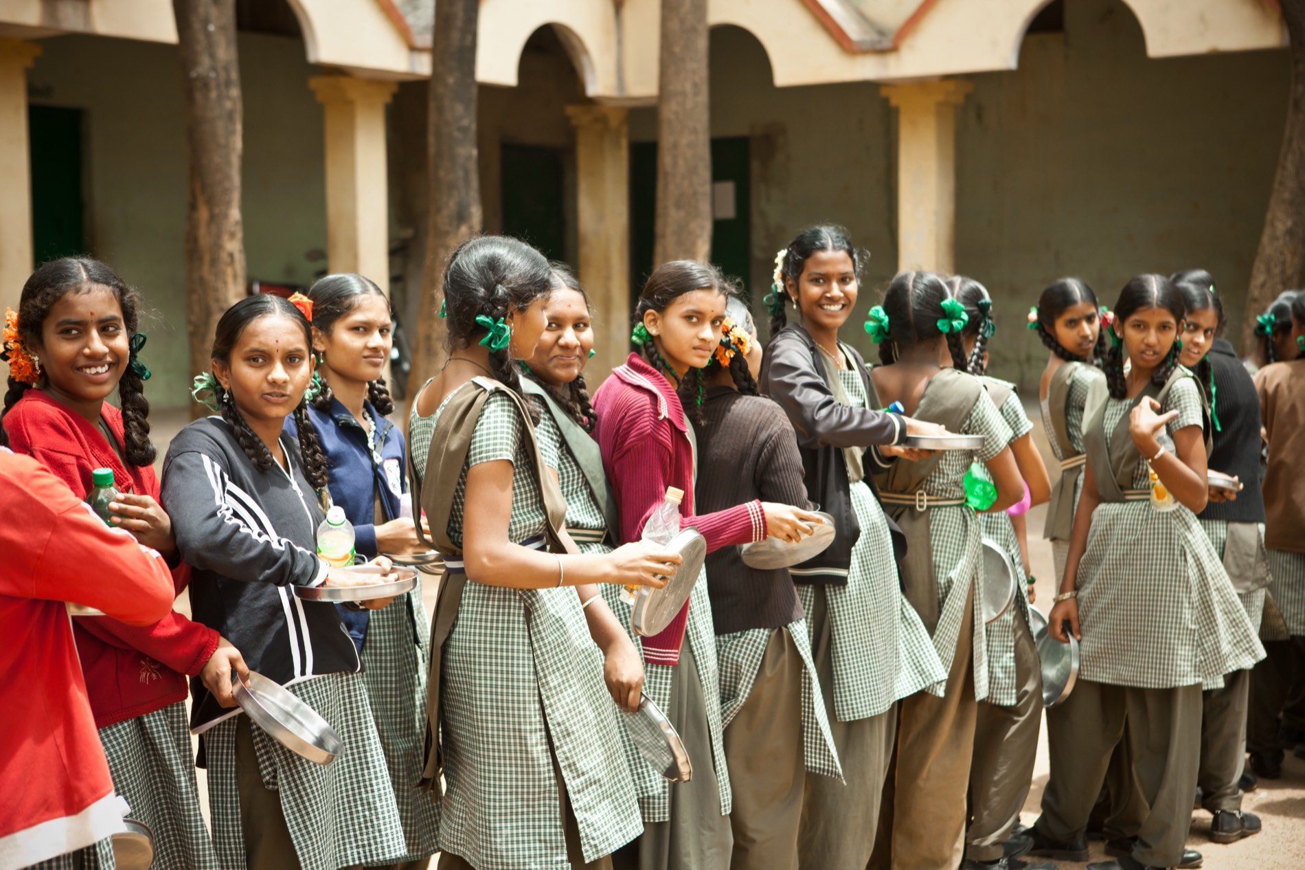 Schoolgirls in green checked uniforms queuing in a courtyard holding steel plates and bowls.