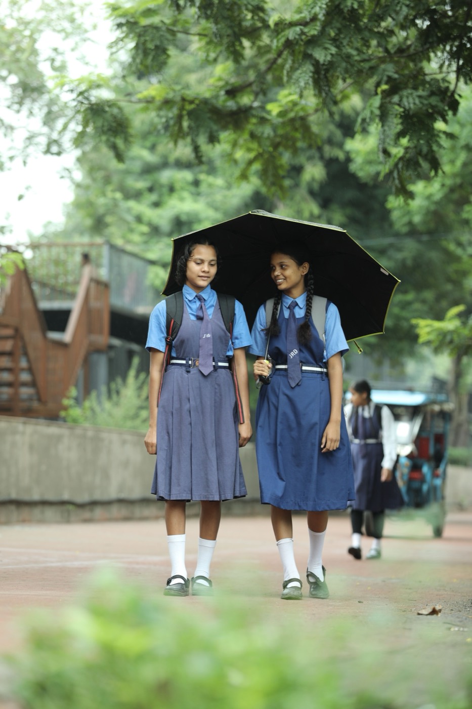 Two schoolgirls in blue uniforms walking together under an umbrella along a tree-lined path.